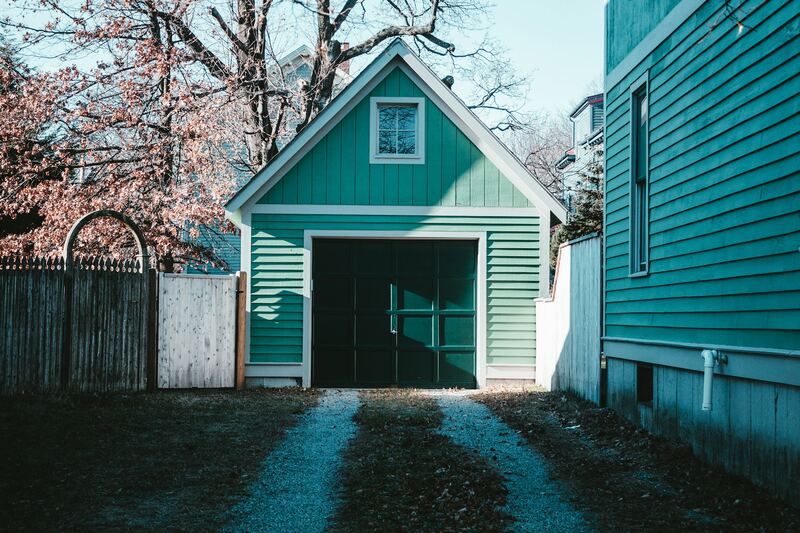 Green carriage-house style detached garage with dark paneled doors beyond a gravel driveway